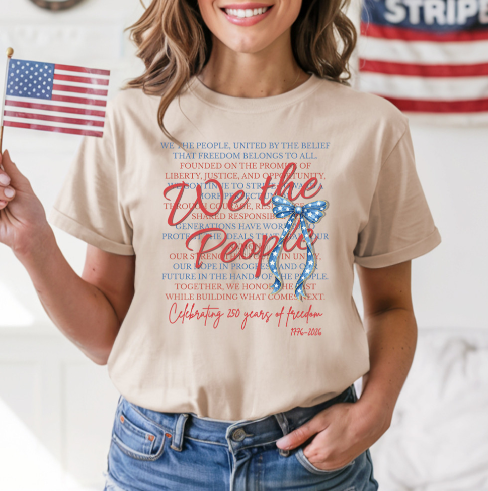 a soft beige unisex Gildan t-shirt with a crew neckline, short sleeves, layered patriotic front wording, a star-spangled bow graphic, a 1776-2026 commemorative detail, and an easy year-round fit