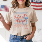 a soft beige unisex Gildan t-shirt with a crew neckline, short sleeves, layered patriotic front wording, a star-spangled bow graphic, a 1776-2026 commemorative detail, and an easy year-round fit