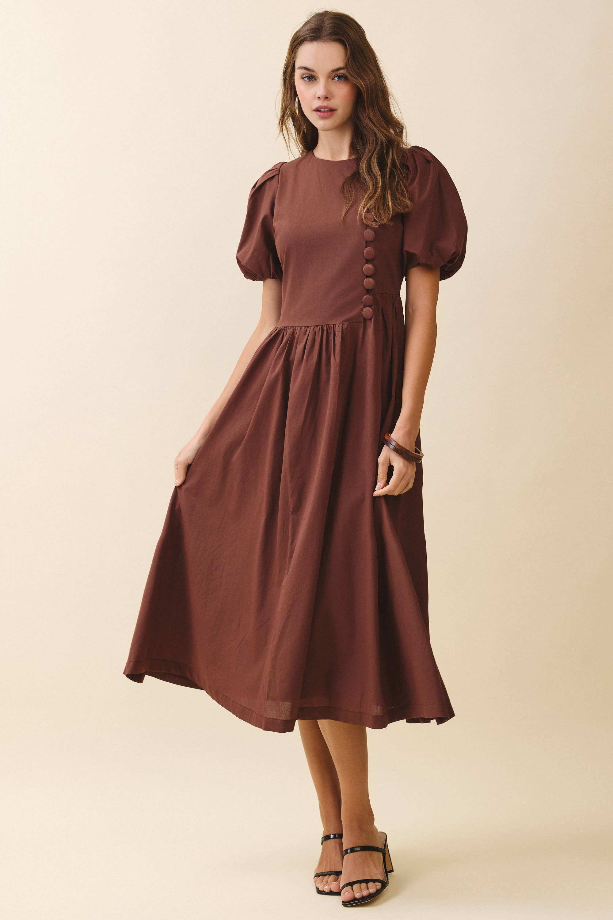 a brown puff sleeve midi dress with a rounded neckline, fitted bodice accented with fabric-covered faux buttons, gathered waist, full A-line skirt, side pockets, and a concealed back zipper