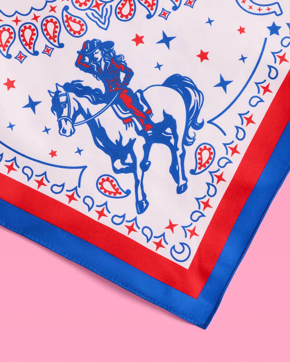 a square bandana with a red, white, and blue western-inspired print, complete with a bold border, cowgirls, hearts, horses, horseshoes, paisley accents, and stars