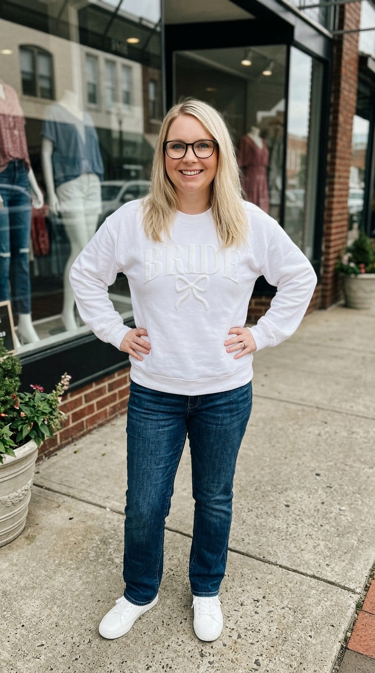 Woman wearing a white sweater and blue jeans standing on a sidewalk with a store in the background.
