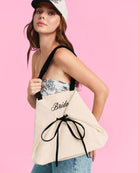 a roomy canvas tote with black embroidered 'Bride' script, black shoulder straps, and bow tie detail