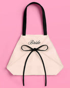 a roomy canvas tote with black embroidered 'Bride' script, black shoulder straps, and bow tie detail