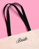 a roomy cream canvas tote with black embroidered 'Bride' script, black shoulder straps, and bow tie detail