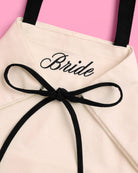 a roomy cream canvas tote with black embroidered 'Bride' script, black shoulder straps, and bow tie detail