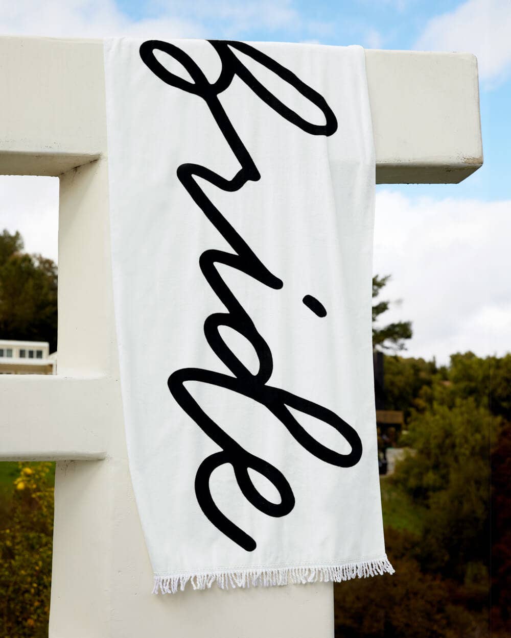White towel with 'Bride' in black script hanging on a white railing outdoors.