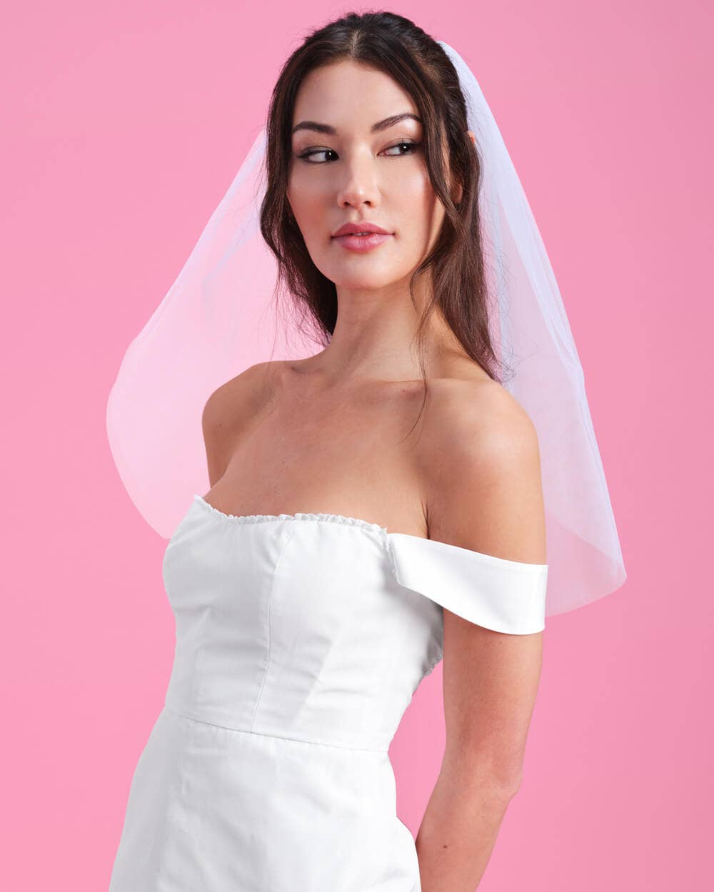 Woman wearing a white off-shoulder dress with a sheer veil against a pink background