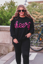 black long sleeve sweater adorned with 'cheers' in pink tinsel 