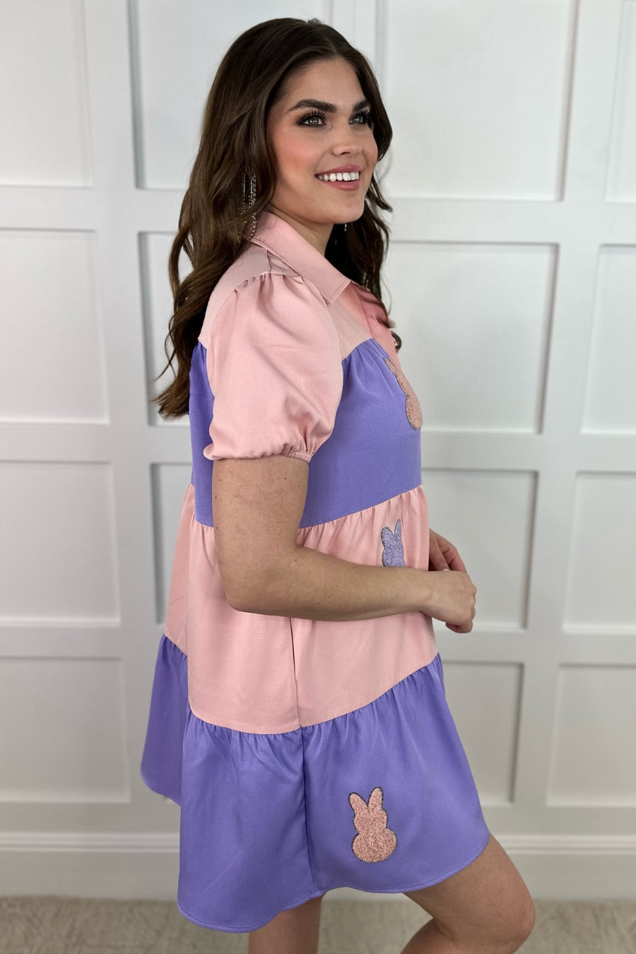 a short-sleeve collared, multi-tiered button-down dress with pockets, designed in a pink and purple vertical color block stripe pattern and finished with alternating Easter bunny patch appliqués