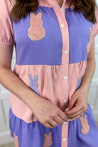 a short-sleeve collared, multi-tiered button-down dress with pockets, designed in a pink and purple vertical color block stripe pattern and finished with alternating Easter bunny patch appliqués