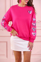 a hot pink long-sleeve pullover made from luxe material with a relaxed drop-shoulder silhouette, a classic crew neckline, and colorful sequin heart accents along the sleeves and chest