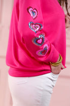 a hot pink long-sleeve pullover made from luxe material with a relaxed drop-shoulder silhouette, a classic crew neckline, and colorful sequin heart accents along the sleeves and chest