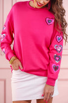 a hot pink long-sleeve pullover made from luxe material with a relaxed drop-shoulder silhouette, a classic crew neckline, and colorful sequin heart accents along the sleeves and chest