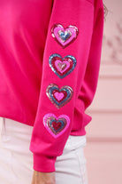 a hot pink long-sleeve pullover made from luxe material with a relaxed drop-shoulder silhouette, a classic crew neckline, and colorful sequin heart accents along the sleeves and chest
