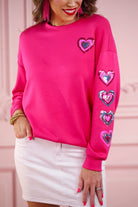a hot pink long-sleeve pullover made from luxe material with a relaxed drop-shoulder silhouette, a classic crew neckline, and colorful sequin heart accents along the sleeves and chest