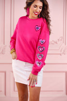 a hot pink long-sleeve pullover made from luxe material with a relaxed drop-shoulder silhouette, a classic crew neckline, and colorful sequin heart accents along the sleeves and chest