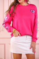 a hot pink long-sleeve pullover made from luxe material with a relaxed drop-shoulder silhouette, a classic crew neckline, and colorful sequin heart accents along the sleeves and chest