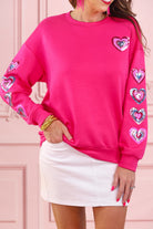a hot pink long-sleeve pullover made from luxe material with a relaxed drop-shoulder silhouette, a classic crew neckline, and colorful sequin heart accents along the sleeves and chest