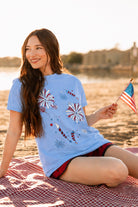 a relaxed blue crew neck tee with a screenprinted fireworks design (rockets, bursts, and starry details), short sleeves, standard t-shirt length, unlined construction, and soft cotton with light stretch