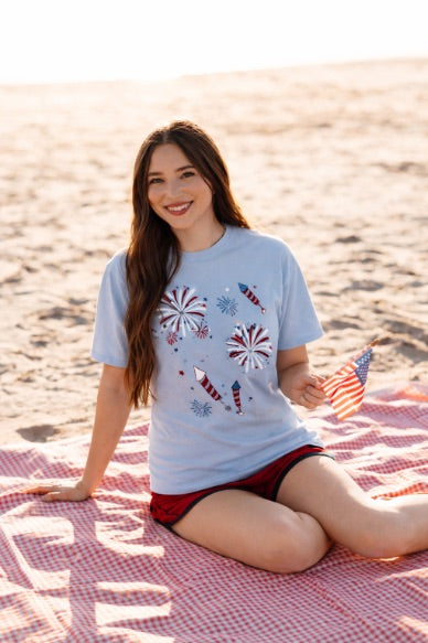 a relaxed blue crew neck tee with a screenprinted fireworks design (rockets, bursts, and starry details), short sleeves, standard t-shirt length, unlined construction, and soft cotton with light stretch