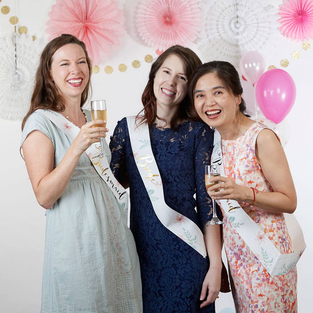 Three women celebrating with champagne glasses and bachelorette sashes in a decorated room.