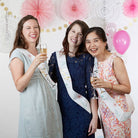 Three women celebrating with champagne glasses and bachelorette sashes in a decorated room.