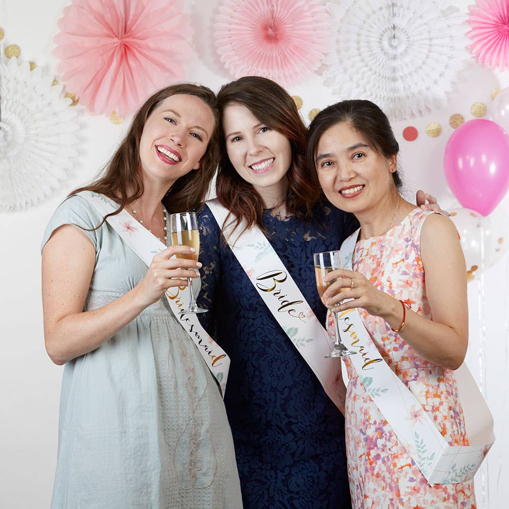 Three women celebrating with champagne at a bachelorette party, wearing sashes and posing against a decorated wall.