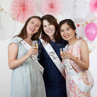 Three women celebrating with champagne at a bachelorette party, wearing sashes and posing against a decorated wall.