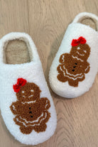 white plush slippers crafted from sherpa style material adorned with an adorable chenille gingerbread girl topped with a festive red bow, a soft interior lining, and easy slip-on silhouette