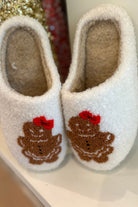 white plush slippers crafted from sherpa style material adorned with an adorable chenille gingerbread girl topped with a festive red bow, a soft interior lining, and easy slip-on silhouette