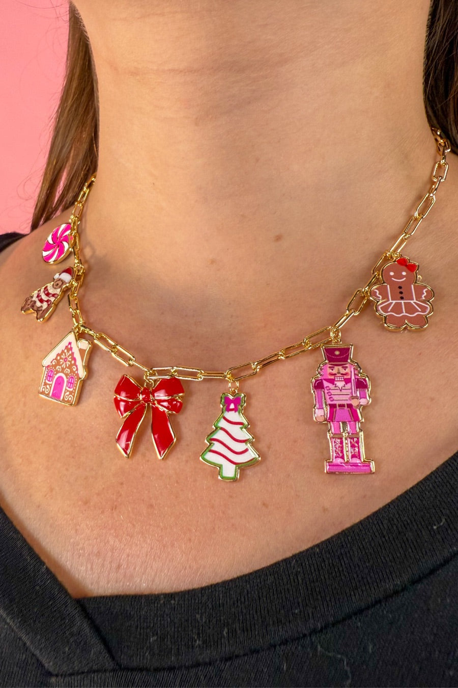 an adjustable gold link paperclip-style necklace adorned with whimsical enamel charms (a bold red bow, Christmas bear, Christmas tree, gingerbread girl, gingerbread house, pink nutcracker, and a pink peppermint)