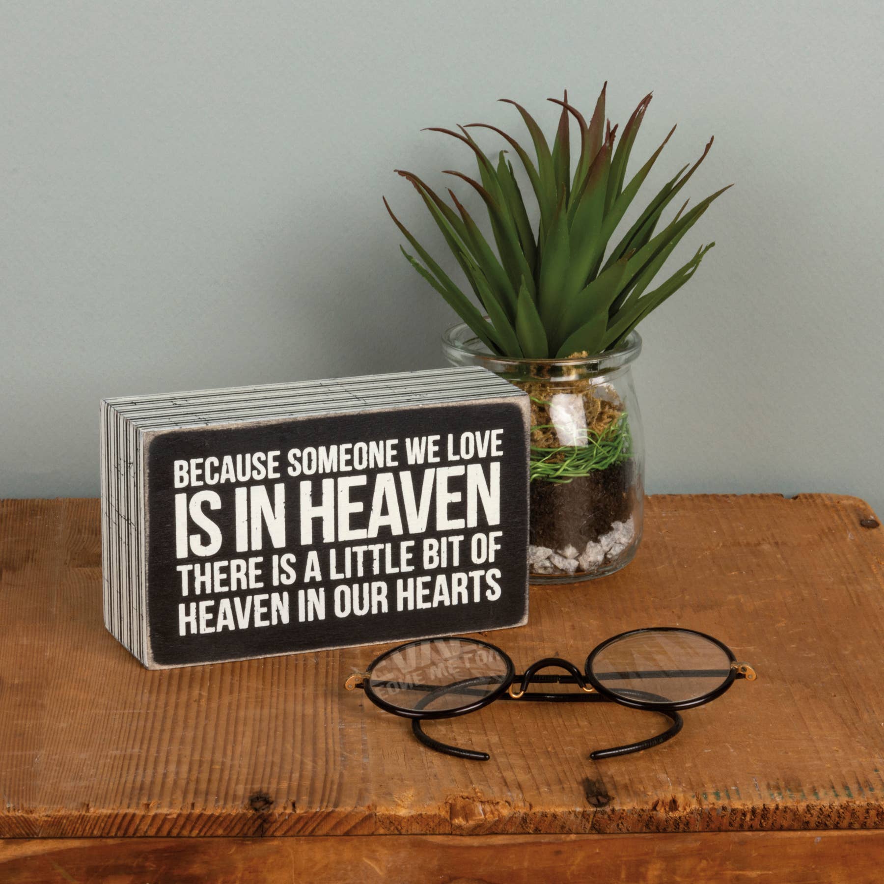 Decorative sign with a quote, glasses, and a plant on a wooden surface.