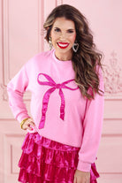 a light pink long-sleeve pullover made from luxe material with drop shoulder, a crew neckline, and a foil bow outlined in delicate pearls