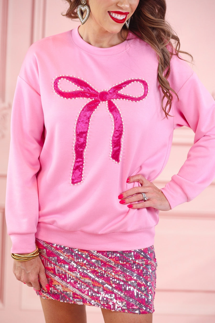 a light pink long-sleeve pullover made from luxe material with drop shoulder, a crew neckline, and a foil bow outlined in delicate pearls