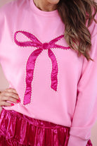 a light pink long-sleeve pullover made from luxe material with drop shoulder, a crew neckline, and a foil bow outlined in delicate pearls