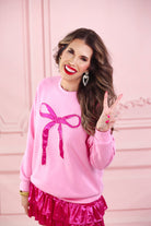 a light pink long-sleeve pullover made from luxe material with drop shoulder, a crew neckline, and a foil bow outlined in delicate pearls