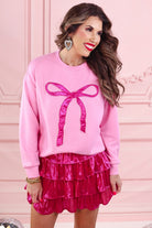 a light pink long-sleeve pullover made from luxe material with drop shoulder, a crew neckline, and a foil bow outlined in delicate pearls