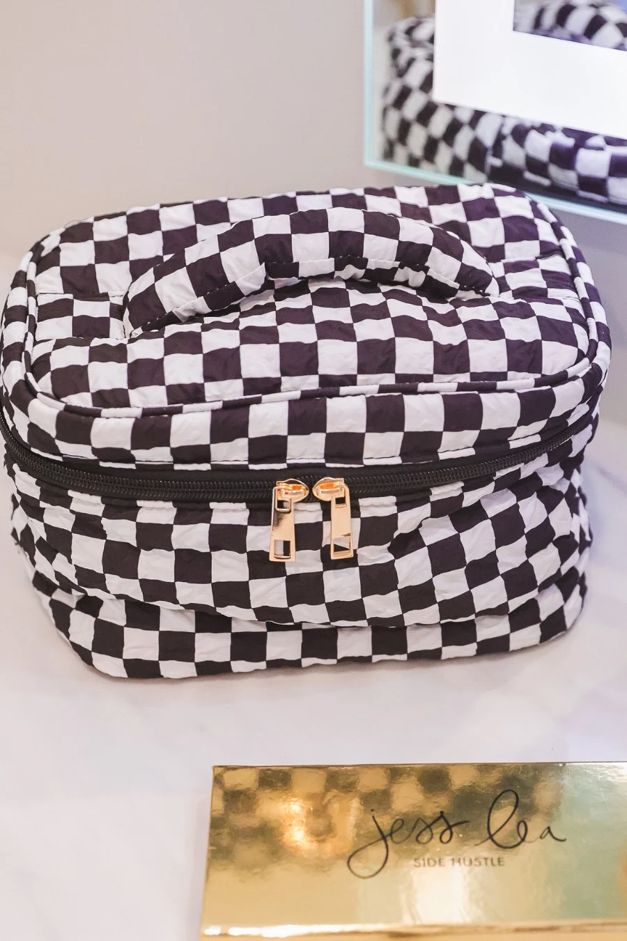 A black and white checkered cosmetic bag with a top handle and dual gold-tone zipper pulls, resting on a light surface next to a gold makeup palette.