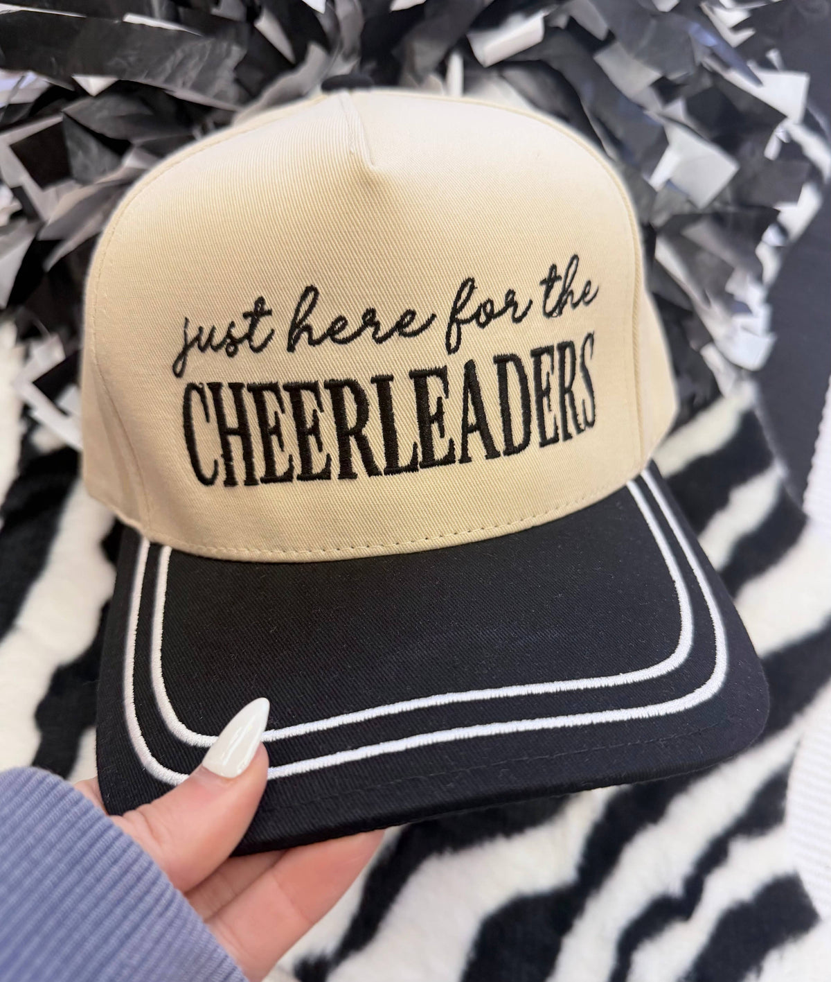 a lightweight unisex embroidered hat with black script 'just here for the' lettering, bold block 'CHEERLEADERS' text, contrasting black bill, white piped trim, cream structured front, black top button, and adjustable snap-back closure