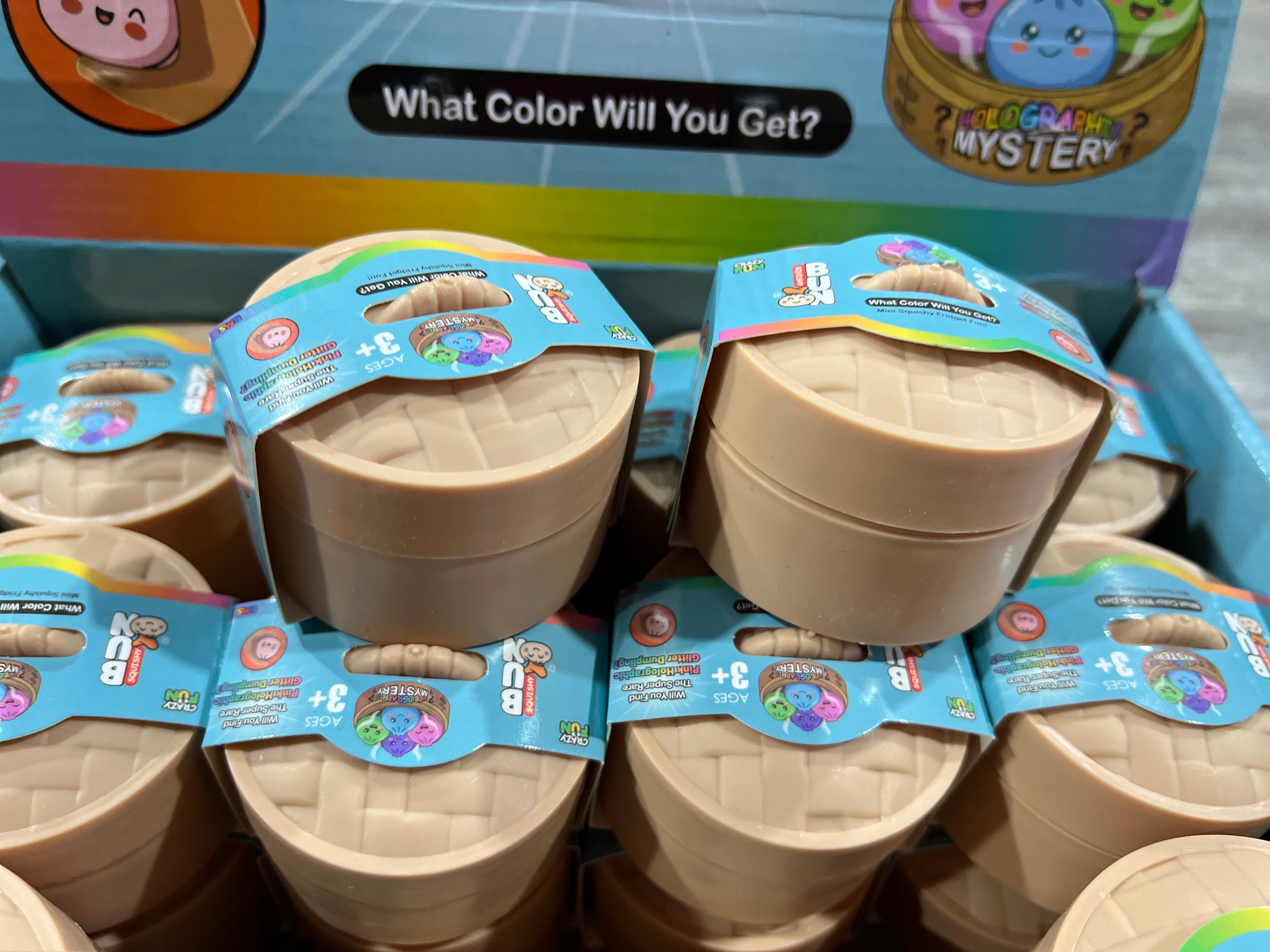A retail display box filled with small round plastic mystery toy containers. The containers are beige/tan with blue and multicolor labels, and the top box reads 'What Color Will You Get?' and 'Mystery'.