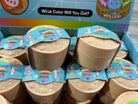 A retail display box filled with small round plastic mystery toy containers. The containers are beige/tan with blue and multicolor labels, and the top box reads 'What Color Will You Get?' and 'Mystery'.