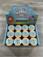 A retail display box of 'The Viral Mystery Dumpling' products, showing twelve individual round packages arranged inside a blue and white cardboard display unit on a gray wood-textured surface.
