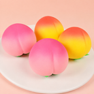peach-shaped squishy toys with a soft touch texture, rounded fruit design, center groove, green leaf accent, and clear individual packaging in pink ombré and yellow-orange ombré