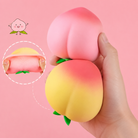 peach-shaped squishy toys with a soft touch texture, rounded fruit design, center groove, green leaf accent, and clear individual packaging in pink ombré and yellow-orange ombré