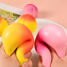 peach-shaped squishy toys with a soft touch texture, rounded fruit design, center groove, green leaf accent, and clear individual packaging in pink ombré and yellow-orange ombré