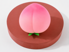 a pink ombré peach-shaped squishy toy with a soft touch texture, rounded fruit design, center groove, green leaf accent, and clear individual packaging