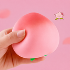 a pink ombré peach-shaped squishy toy with a soft touch texture, rounded fruit design, center groove, green leaf accent, and clear individual packaging