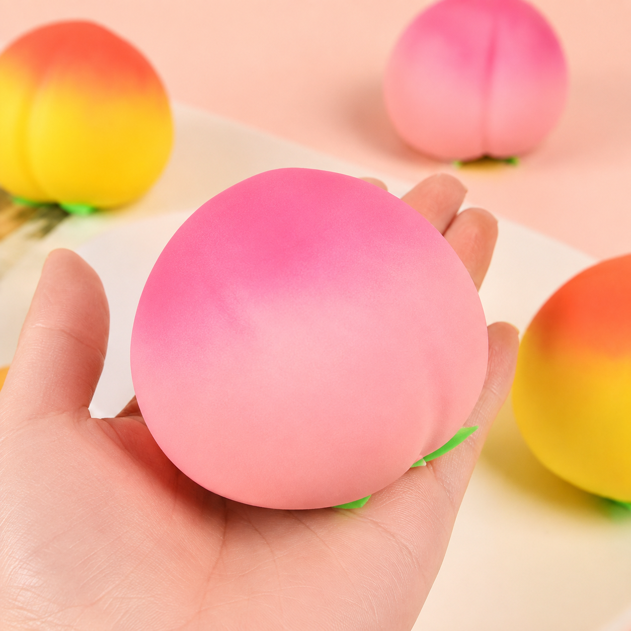 a pink ombré peach-shaped squishy toy with a soft touch texture, rounded fruit design, center groove, green leaf accent, and clear individual packaging