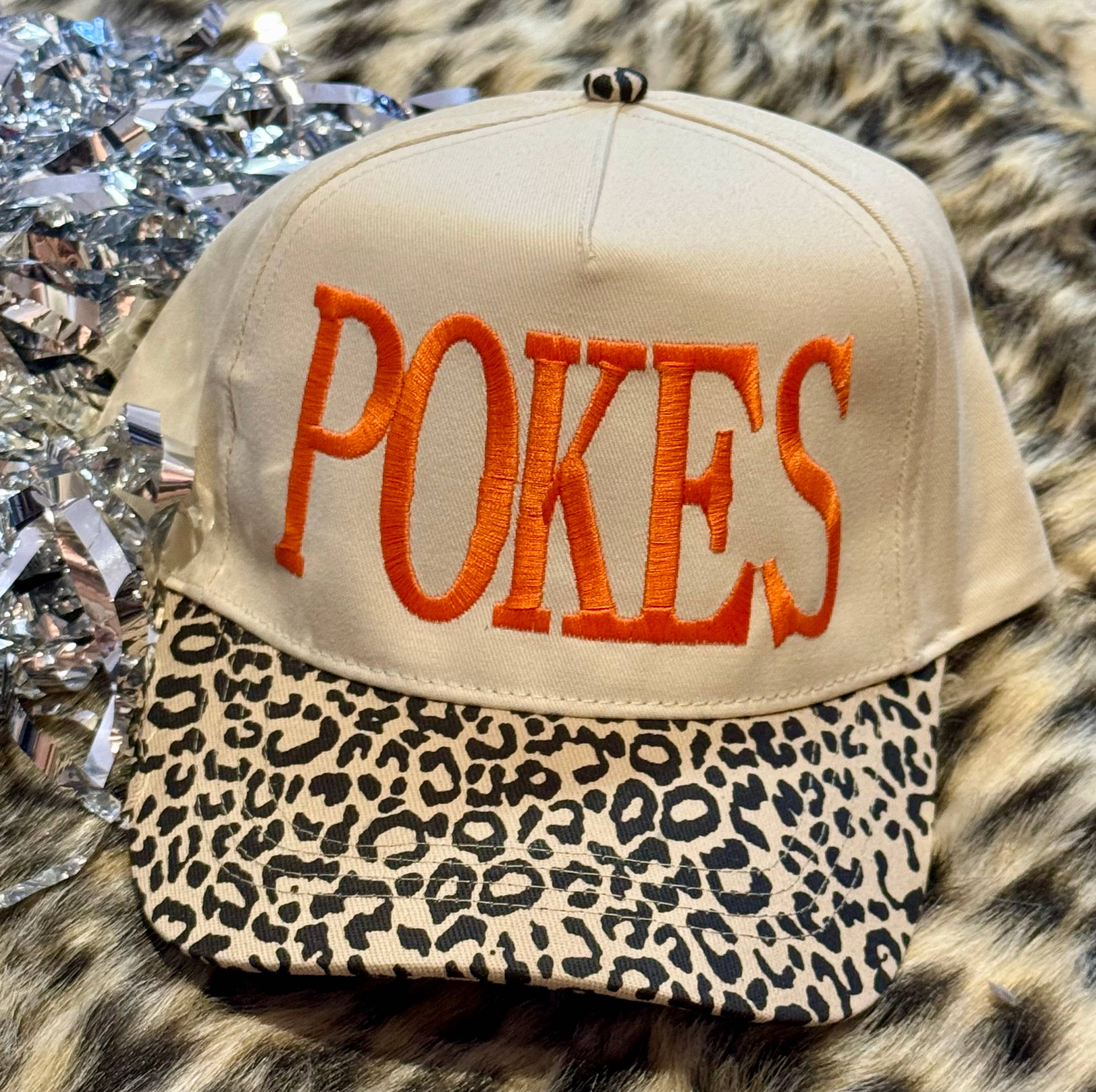 a lightweight unisex two-tone hat with embroidered orange 'POKES' lettering, cream structured front, curved contrasting black leopard bill, coordinating leopard top button, and adjustable snap-back closure
