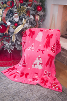 A bright pink fleece throw blanket with a red and white Christmas pattern featuring bows, trees, and ornaments, draped over a chair in a living room setting next to a decorated Christmas tree.